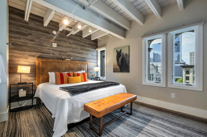 Modern bedroom with a large bed, wooden accents, white linens, and city view windows.