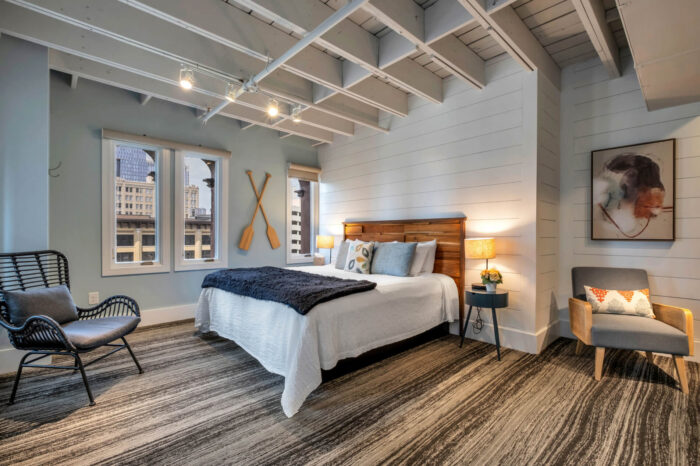 Modern bedroom with exposed beams, city view windows, plush bedding, and artistic decor.