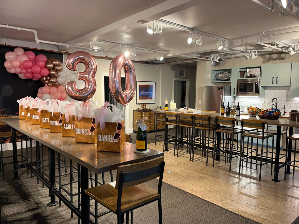 Room decorated for a 30th birthday with balloons, gift bags on a table, and a kitchen area in the background.