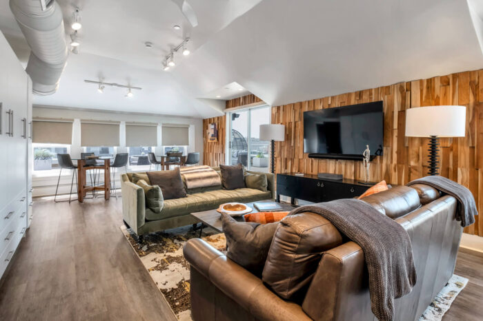 Modern living room with brown sofas, wooden wall paneling, and white kitchen in the background.