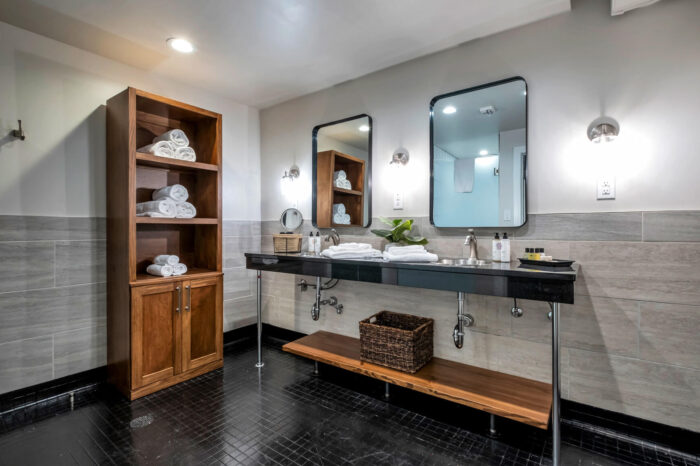 Modern bathroom with double sink, large mirrors, wooden shelves, and grey tiles.