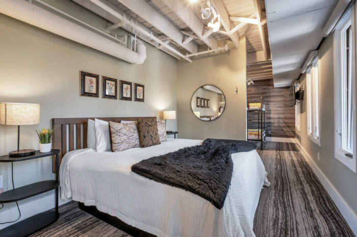 Modern bedroom with exposed pipes, wood accents, large bed with decorative pillows, and circular mirror.