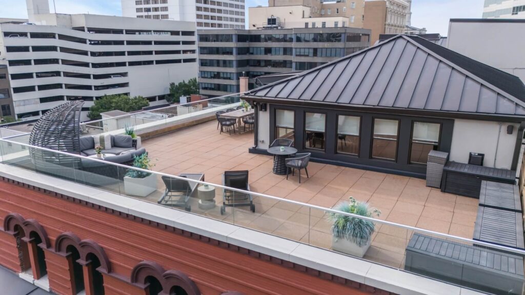Rooftop patio with furniture and glass railing, overlooking urban buildings.