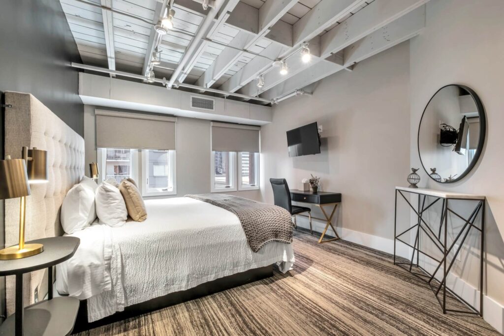 Modern bedroom with a bed, side table, lamp, desk, chair, TV, and exposed ceiling beams.