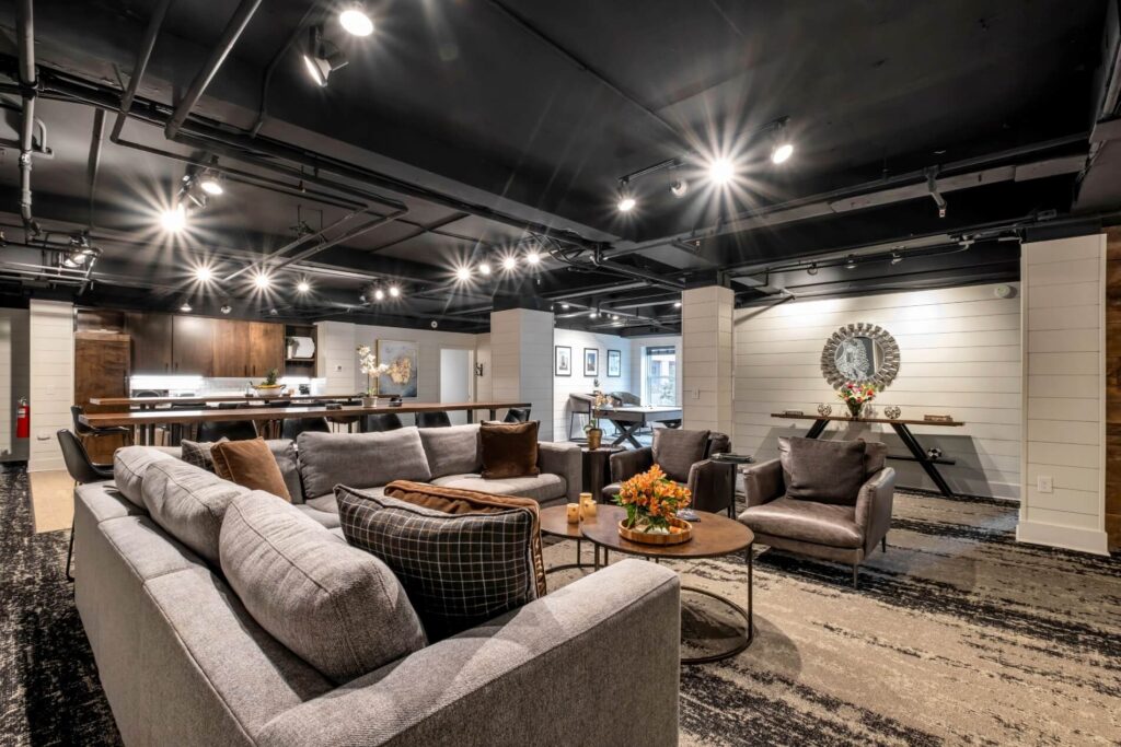 Modern living room with sectional sofa, kitchen area, exposed ceiling pipes, and warm lighting.
