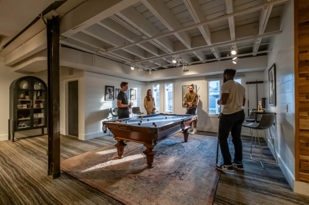 People playing pool in a cozy room with white walls and wooden beams.