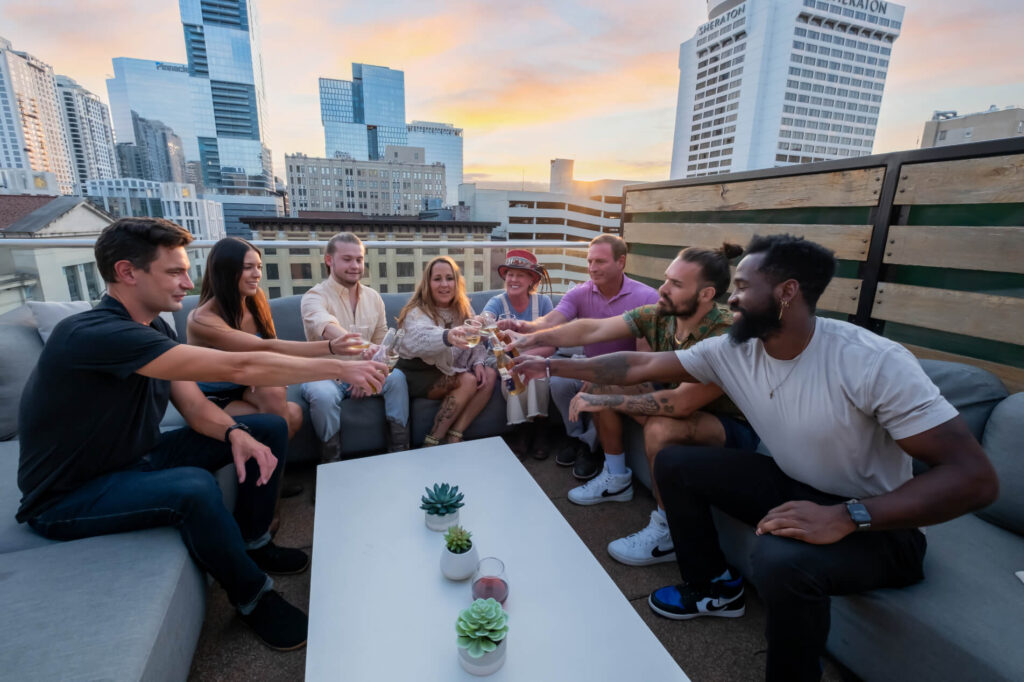 Group of friends toasting drinks on a rooftop at sunset with city skyline in the background.