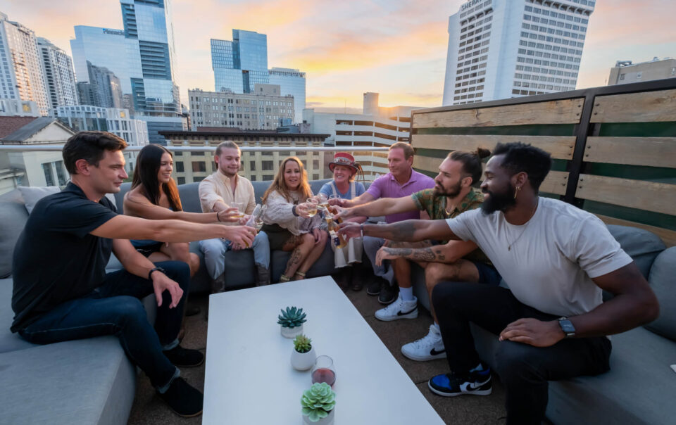 Group of friends toasting drinks on a rooftop at sunset with city skyline in the background.