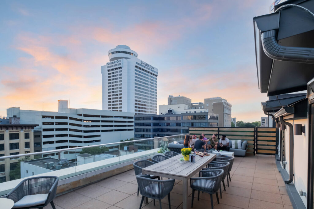 Rooftop patio with people and outdoor furniture overlooking urban skyline at dusk.
