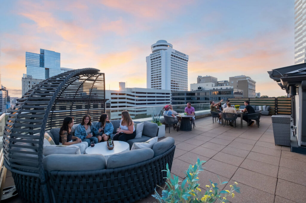 Groups of people socializing on a rooftop patio at dusk with city skyline in the background.