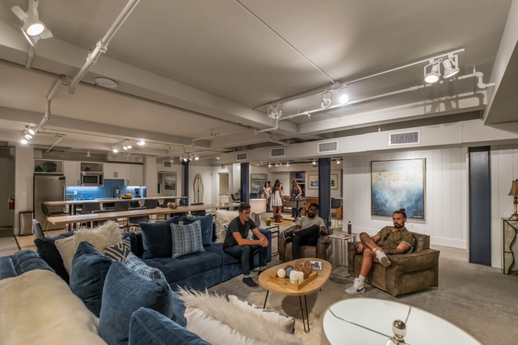 Spacious living area with group of people, modern furniture, kitchen in background.