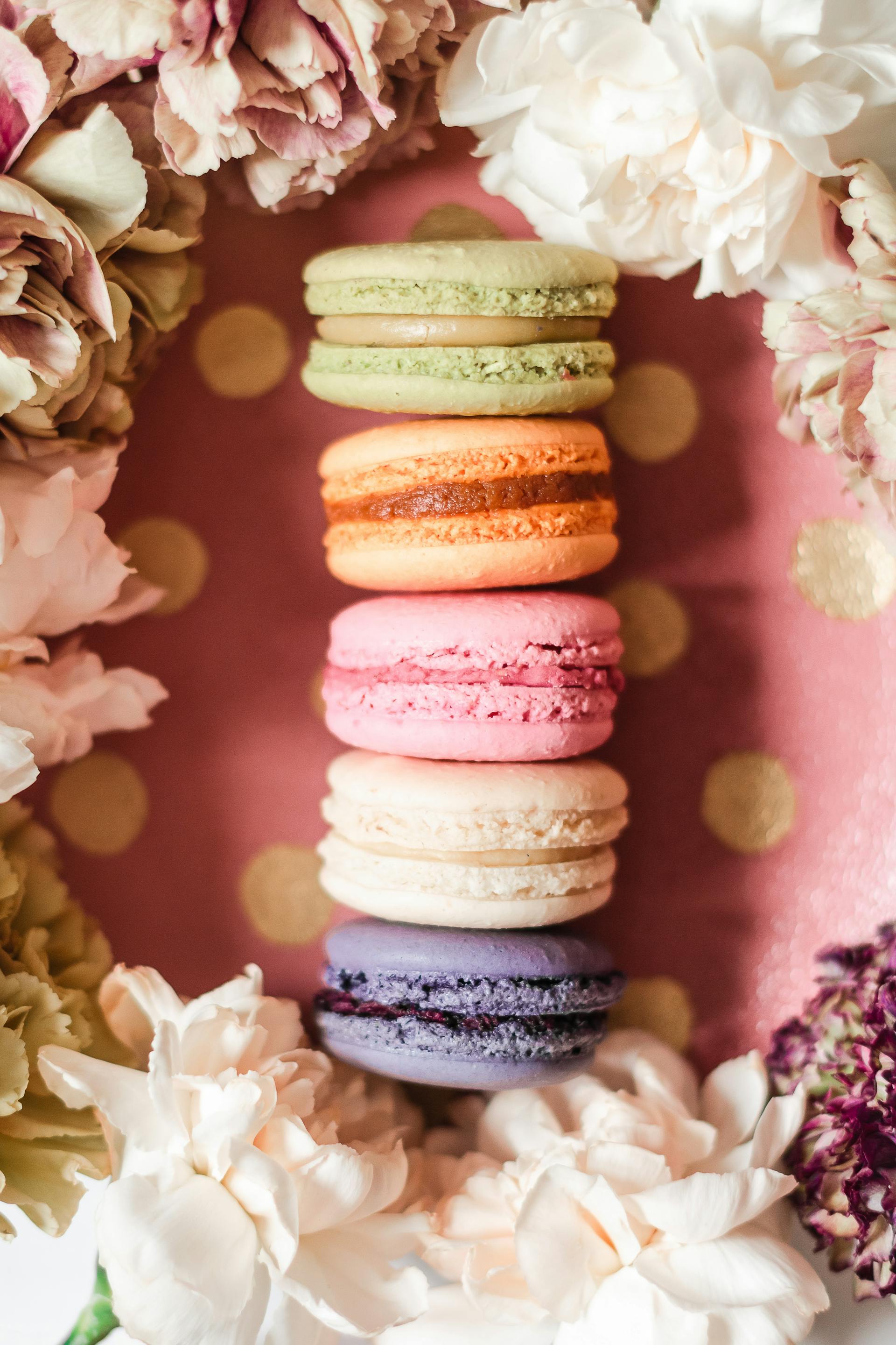 a stack of colorful macarons surrounded by flowers on a pink polka dot background