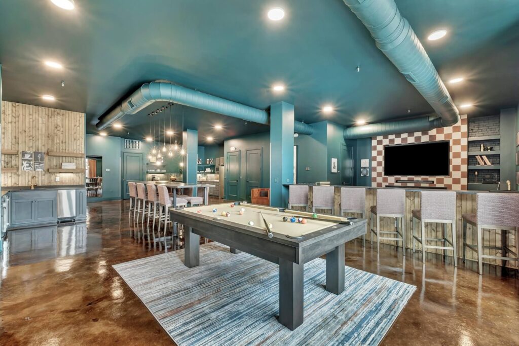 A modern game room or common area with a teal blue ceiling, exposed ductwork, and polished concrete floors. The room features a pool table with cues and balls, a bar area with high stools and a large flat-screen TV mounted on a checkered wall, and a dining or lounge area with a long table and upholstered chairs. There's also a kitchen or serving area with wooden paneling and stainless steel appliances.