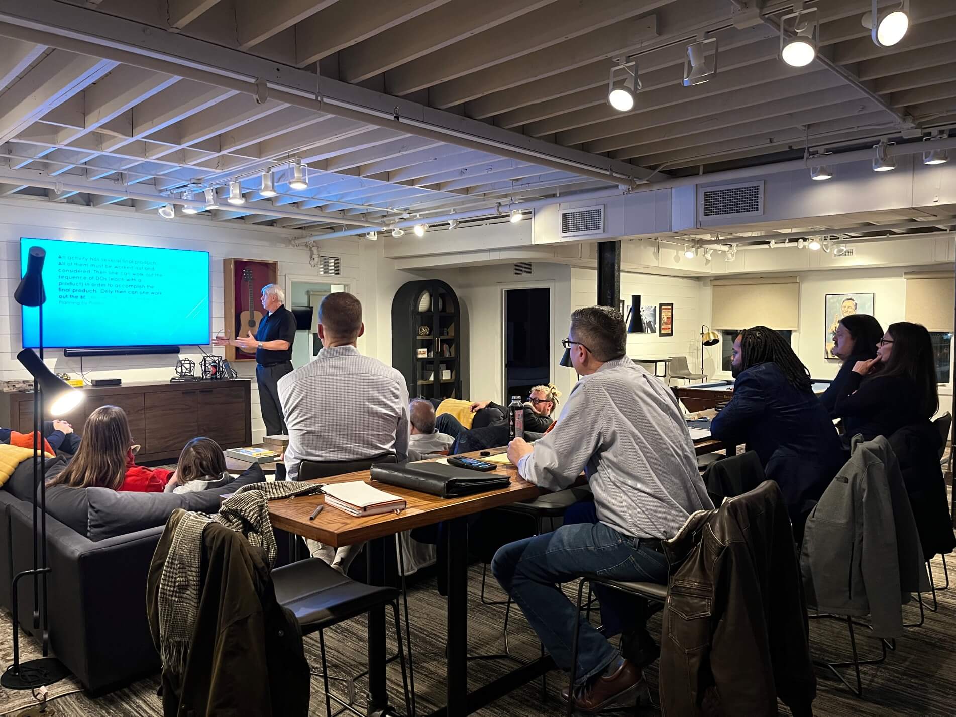 A group of people sitting around tables in a meeting room with a projector screen.